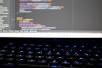 A close-up view of a computer screen displaying lines of colorful programming code. The keyboard in the foreground is slightly blurred, with keys lit by blue backlighting. The code on the screen includes syntax highlighting with various colors used to differentiate between components.