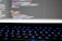 A close-up view of a computer screen displaying lines of colorful programming code. The keyboard in the foreground is slightly blurred, with keys lit by blue backlighting. The code on the screen includes syntax highlighting with various colors used to differentiate between components.