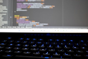 A close-up view of a computer screen displaying lines of colorful programming code. The keyboard in the foreground is slightly blurred, with keys lit by blue backlighting. The code on the screen includes syntax highlighting with various colors used to differentiate between components.