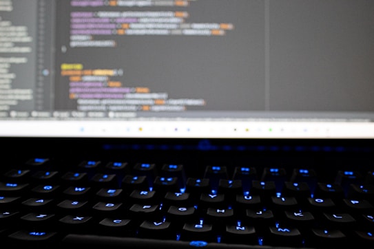 A close-up view of a computer screen displaying lines of colorful programming code. The keyboard in the foreground is slightly blurred, with keys lit by blue backlighting. The code on the screen includes syntax highlighting with various colors used to differentiate between components.