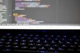 A close-up view of a computer screen displaying lines of colorful programming code. The keyboard in the foreground is slightly blurred, with keys lit by blue backlighting. The code on the screen includes syntax highlighting with various colors used to differentiate between components.