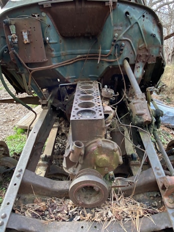 An old, rusted engine block sits exposed on a vehicle chassis in an outdoor setting. Surrounding debris and overgrown vegetation suggest abandonment and decay. The metallic components show significant signs of degradation and corrosion, with various cables and pipes scattered around the structure.