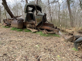 An old, rusted truck chassis lies abandoned in a wooded area, surrounded by leafless trees and dry grass. The body is missing parts, revealing the worn metal and decaying structure.