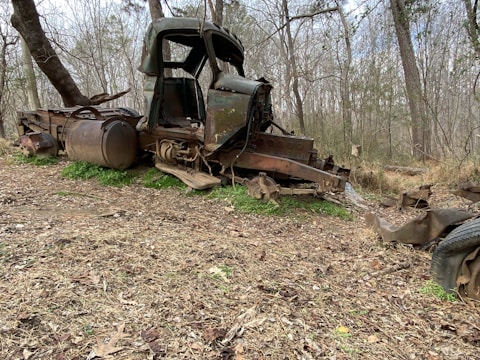 An old, rusted truck chassis lies abandoned in a wooded area, surrounded by leafless trees and dry grass. The body is missing parts, revealing the worn metal and decaying structure.