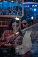 A young couple seated in a cozy indoor setting, with a brick wall and a comic-style poster behind them. The woman is wearing glasses and a red leopard print top, focused on a smartphone she is holding. The man leans in closely beside her, with out-of-focus lights reflecting against a window in the foreground, creating a dreamy ambiance.