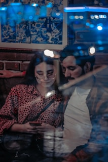 A young couple seated in a cozy indoor setting, with a brick wall and a comic-style poster behind them. The woman is wearing glasses and a red leopard print top, focused on a smartphone she is holding. The man leans in closely beside her, with out-of-focus lights reflecting against a window in the foreground, creating a dreamy ambiance.