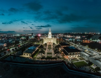 An illuminated church stands prominently in the center of a plaza at night, surrounded by palm trees and gardens. City lights stretch across the horizon under a cloudy blue evening sky.