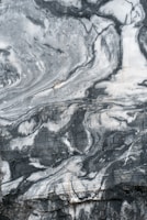 A close-up view of a marble surface showcasing intricate patterns and veins in shades of gray and white. The natural swirls and textures create a dramatic and elegant appearance.