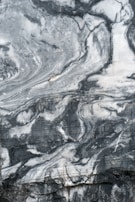 A close-up view of a marble surface showcasing intricate patterns and veins in shades of gray and white. The natural swirls and textures create a dramatic and elegant appearance.