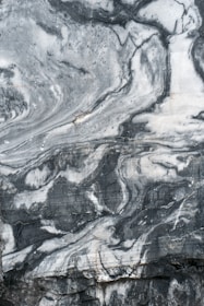 A close-up view of a marble surface showcasing intricate patterns and veins in shades of gray and white. The natural swirls and textures create a dramatic and elegant appearance.