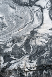 A close-up view of a marble surface showcasing intricate patterns and veins in shades of gray and white. The natural swirls and textures create a dramatic and elegant appearance.