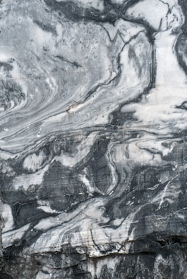 A close-up view of a marble surface showcasing intricate patterns and veins in shades of gray and white. The natural swirls and textures create a dramatic and elegant appearance.
