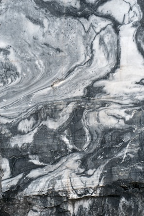 A close-up view of a marble surface showcasing intricate patterns and veins in shades of gray and white. The natural swirls and textures create a dramatic and elegant appearance.