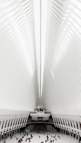An expansive interior space featuring a high, ribbed ceiling with a symmetrical design. The architecture is modern and futuristic, with a bright, white color scheme. People can be seen scattered across the vast floor, highlighting the scale of the structure.