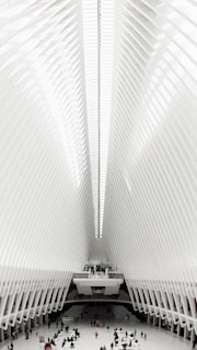 An expansive interior space featuring a high, ribbed ceiling with a symmetrical design. The architecture is modern and futuristic, with a bright, white color scheme. People can be seen scattered across the vast floor, highlighting the scale of the structure.