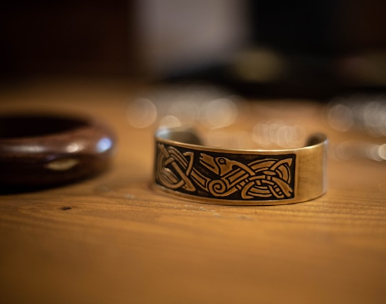 A close-up of a metal bracelet with intricate Celtic designs etched onto its surface. The bracelet rests on a wooden surface, and the background is softly blurred, giving a warm, intimate atmosphere.