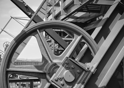 A close-up view of a large industrial metal wheel with visible beams and structural components. The machinery appears robust and intricately designed, positioned against a bright sky. The metal surfaces have a textured appearance, and the structure includes several other gears and bolts.