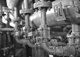 Close-up view of a network of industrial pipes and valves with a monochrome color scheme. The metal surfaces appear aged and somewhat corroded, with visible bolts and joints. The numbering on the pipes indicates some sort of system organization or identification.