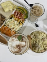 A table set with a variety of dishes including a creamy pasta with bread, a vibrant salad featuring shredded carrots, cabbage, corn, chopped tomatoes, and avocados, alongside a folded omelet and piece of flatbread. Two drinks are present: one is a tall glass of iced coffee with visible ice cubes, and the other appears to be a cappuccino with frothy milk and a mint leaf garnish.