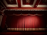 A theater stage is adorned with rich, red velvet curtains that are drawn closed. The gilt and ornate decor around the stage, including an intricately designed ceiling, enhances the luxurious appearance of the setting.