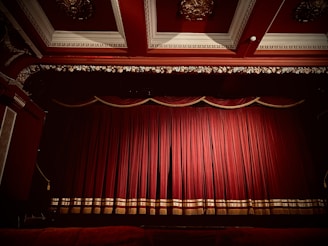 A theater stage is adorned with rich, red velvet curtains that are drawn closed. The gilt and ornate decor around the stage, including an intricately designed ceiling, enhances the luxurious appearance of the setting.
