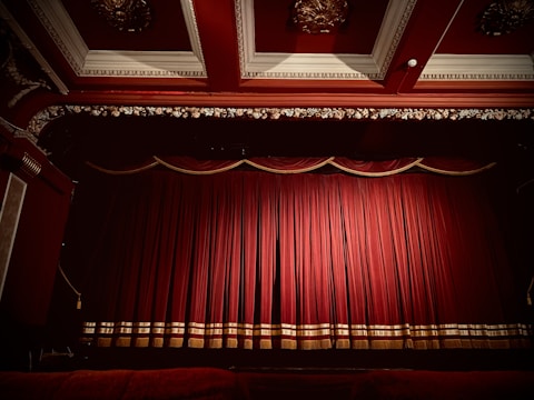 A theater stage is adorned with rich, red velvet curtains that are drawn closed. The gilt and ornate decor around the stage, including an intricately designed ceiling, enhances the luxurious appearance of the setting.