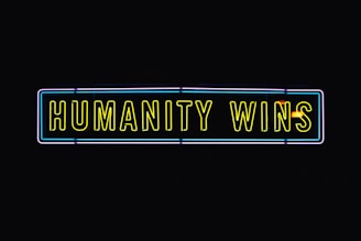A neon sign with the text 'HUMANITY WINS' in bright yellow letters on a black background. The sign has a rectangular shape and an outer neon border with hints of blue and purple.