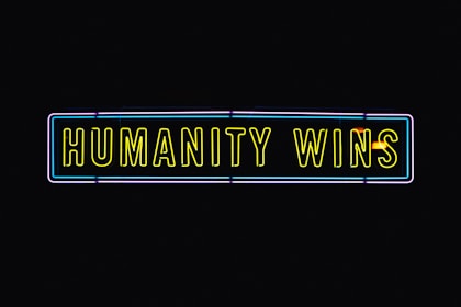 A neon sign with the text 'HUMANITY WINS' in bright yellow letters on a black background. The sign has a rectangular shape and an outer neon border with hints of blue and purple.