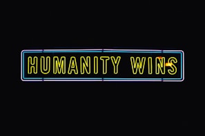 A neon sign with the text 'HUMANITY WINS' in bright yellow letters on a black background. The sign has a rectangular shape and an outer neon border with hints of blue and purple.