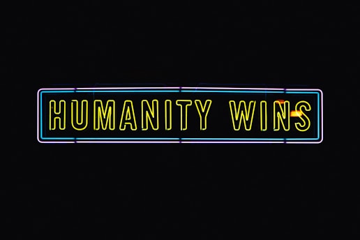 A neon sign with the text 'HUMANITY WINS' in bright yellow letters on a black background. The sign has a rectangular shape and an outer neon border with hints of blue and purple.