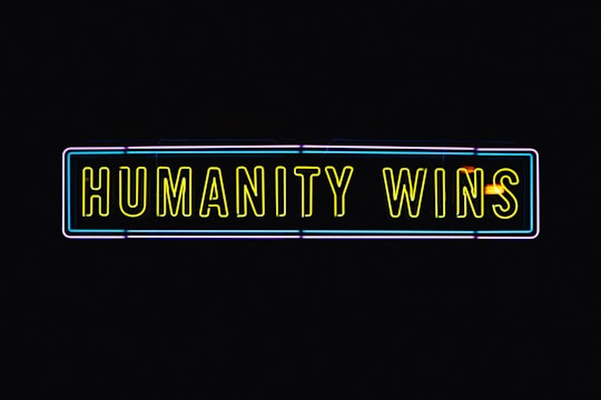 A neon sign with the text 'HUMANITY WINS' in bright yellow letters on a black background. The sign has a rectangular shape and an outer neon border with hints of blue and purple.