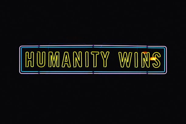 A neon sign with the text 'HUMANITY WINS' in bright yellow letters on a black background. The sign has a rectangular shape and an outer neon border with hints of blue and purple.