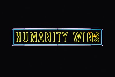 A neon sign with the text 'HUMANITY WINS' in bright yellow letters on a black background. The sign has a rectangular shape and an outer neon border with hints of blue and purple.