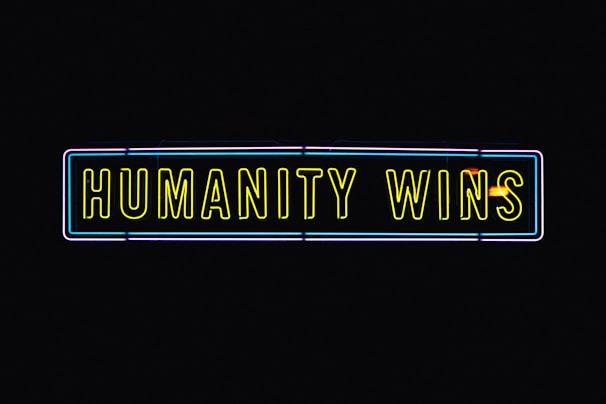 A neon sign with the text 'HUMANITY WINS' in bright yellow letters on a black background. The sign has a rectangular shape and an outer neon border with hints of blue and purple.