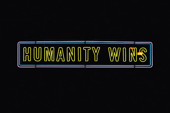 A neon sign with the text 'HUMANITY WINS' in bright yellow letters on a black background. The sign has a rectangular shape and an outer neon border with hints of blue and purple.