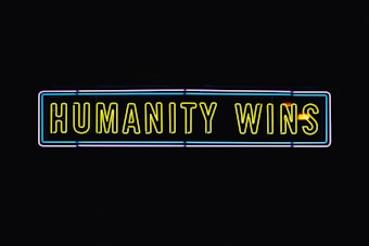 A neon sign with the text 'HUMANITY WINS' in bright yellow letters on a black background. The sign has a rectangular shape and an outer neon border with hints of blue and purple.