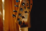 A close-up of a guitar headstock with tuning pegs and strings visible. The brand name and logos are etched on the wooden surface. The background is softly blurred, creating a warm, intimate atmosphere.