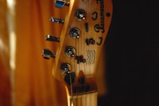 A close-up of a guitar headstock with tuning pegs and strings visible. The brand name and logos are etched on the wooden surface. The background is softly blurred, creating a warm, intimate atmosphere.