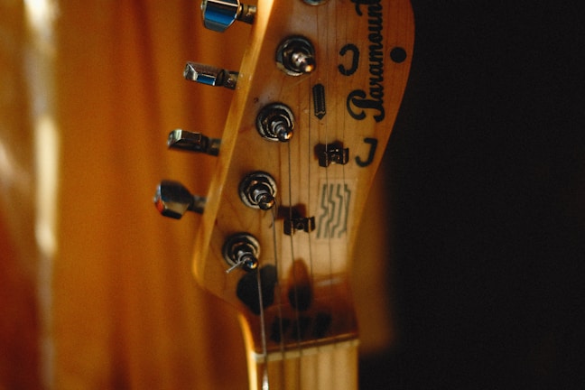 A close-up of a guitar headstock with tuning pegs and strings visible. The brand name and logos are etched on the wooden surface. The background is softly blurred, creating a warm, intimate atmosphere.