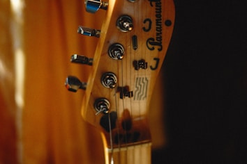 A close-up of a guitar headstock with tuning pegs and strings visible. The brand name and logos are etched on the wooden surface. The background is softly blurred, creating a warm, intimate atmosphere.