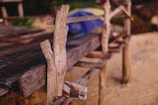 A rustic, weathered wooden structure is partially visible, with rough, natural planks and beams showcasing a handmade or aged quality. The background includes a blurred view of sand and hints of greenery, suggesting an outdoor setting. The overall composition is simple yet textured, emphasizing the natural wear of the wood.