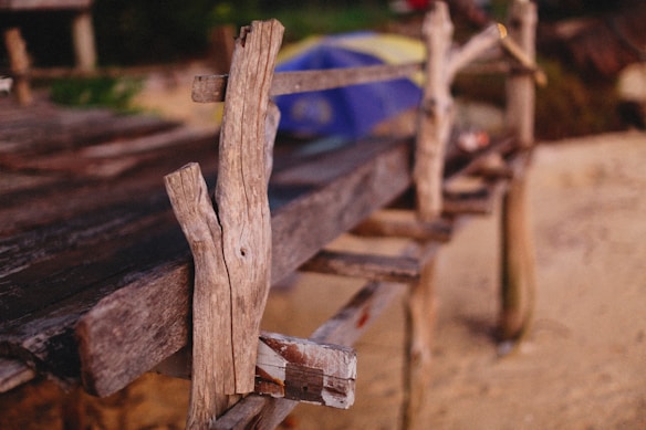 A rustic, weathered wooden structure is partially visible, with rough, natural planks and beams showcasing a handmade or aged quality. The background includes a blurred view of sand and hints of greenery, suggesting an outdoor setting. The overall composition is simple yet textured, emphasizing the natural wear of the wood.