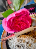 A vibrant pink rose with green leaves placed beside a rolled paper item marked 'HIGHONLOVE'. The setting includes a bundle of dried herbs, possibly sage, resting on a stone surface. In the background, blurred elements and colors add depth.