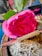 A vibrant pink rose with green leaves placed beside a rolled paper item marked 'HIGHONLOVE'. The setting includes a bundle of dried herbs, possibly sage, resting on a stone surface. In the background, blurred elements and colors add depth.