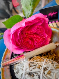 A vibrant pink rose with green leaves placed beside a rolled paper item marked 'HIGHONLOVE'. The setting includes a bundle of dried herbs, possibly sage, resting on a stone surface. In the background, blurred elements and colors add depth.