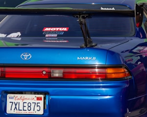 A close-up view of the rear section of a blue Toyota Mark II car. The vehicle has a prominent spoiler and visible branding and license plate from California. The rear window features several decals.