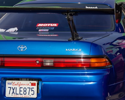 A close-up view of the rear section of a blue Toyota Mark II car. The vehicle has a prominent spoiler and visible branding and license plate from California. The rear window features several decals.