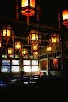 A dimly lit interior with traditional lanterns hanging from the ceiling, casting a warm glow. The room has large decorative windows and dark wooden tables and chairs, giving it an intimate, cozy atmosphere.