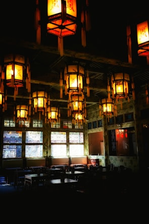 A dimly lit interior with traditional lanterns hanging from the ceiling, casting a warm glow. The room has large decorative windows and dark wooden tables and chairs, giving it an intimate, cozy atmosphere.