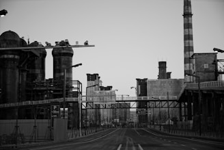 An industrial landscape with large cylindrical storage tanks, a tall chimney, and multiple industrial buildings connected by walkways. The scene is devoid of people, creating a sense of emptiness.